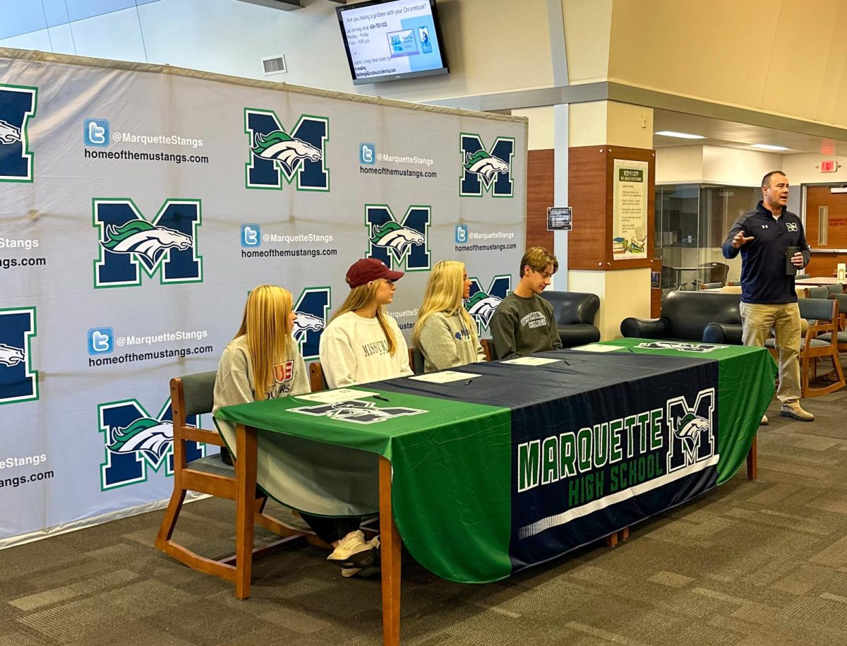 Activities Director Adam Starling commences National Letter of Intent (NLI) signing celebrations before families, friends and coaches in the library on Monday, Nov. 27. From left to right, seniors Carle Bachman, Bri Kappeler, Katie Morgenthaler and Colyn Wright sit with mock NLI contracts.
