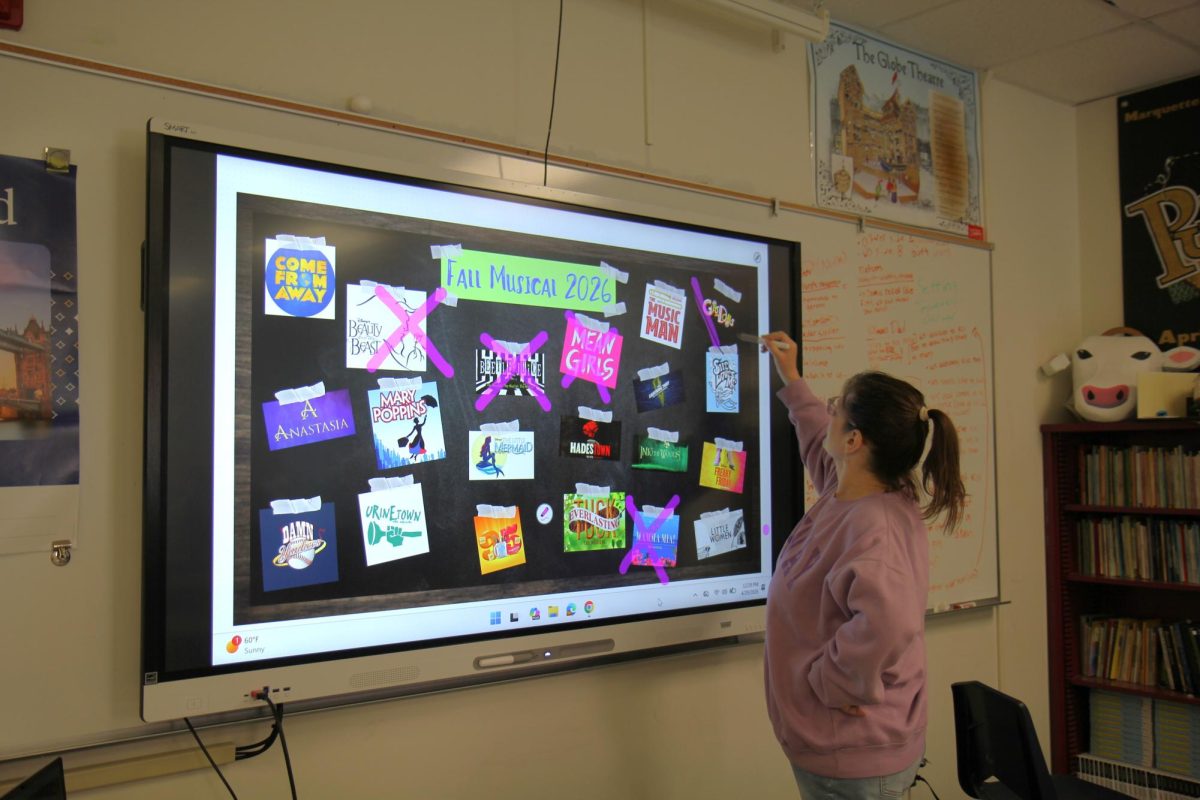 Rebecca Young, theater teacher, stands in front of the possibilities for next year's musical and crosses out the musical "Guys and Dolls" on Wednesday, April 29. Almost every year Young announces the musical for the next year by crossing off options until only one is left.